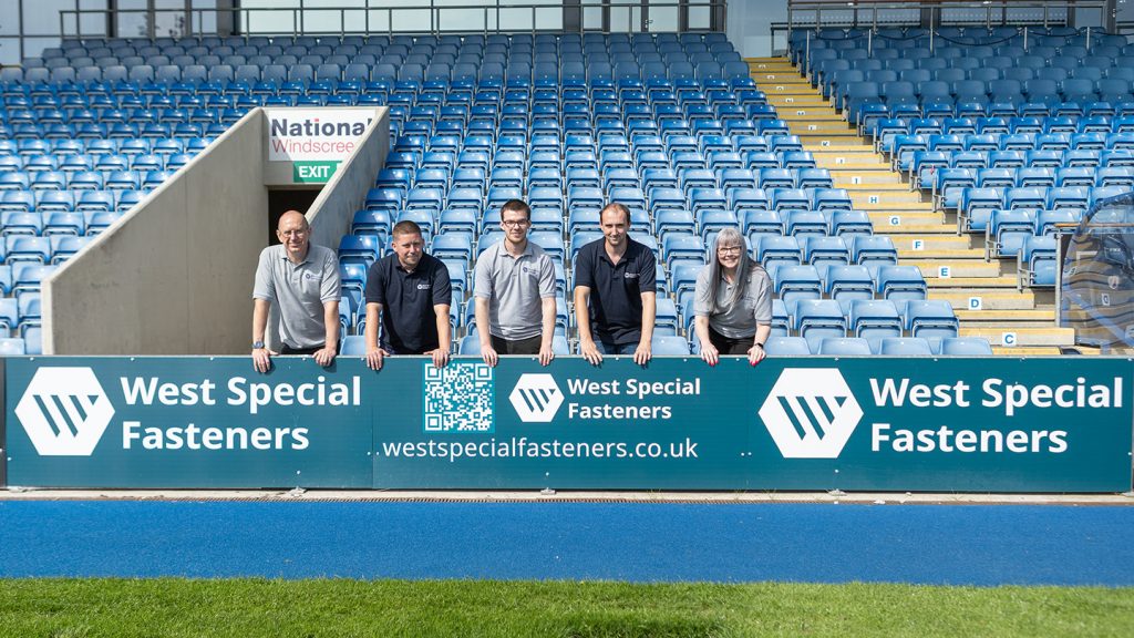 4 colleagues stand in front of the advertising board for West Special Fasteners at Chesterfield Football Club ground